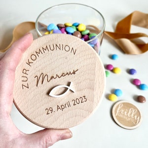 May include: A wooden lid for a jar with the text "Zur Kommunion Marcus 29. April 2023" and a small white fish symbol.