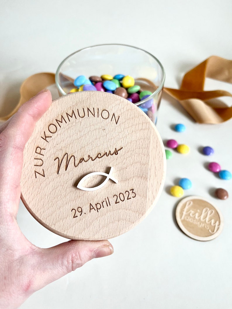 May include: A wooden lid for a jar with the text "Zur Kommunion Marcus 29. April 2023" and a small white fish symbol.