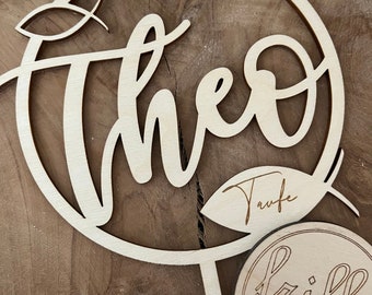 Könnte beinhalten: Holz-Tortenaufsatz mit dem Namen "Theo" in einer kursiv geschriebenen Schrift, umgeben von Blättern. Der Aufsatz enthält auch das Wort "Taufe" und einen kleinen Kreis mit dem Text "frilly designs".