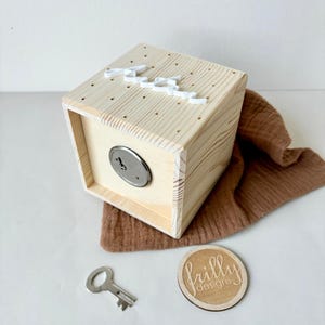 May include: A wooden box with a keyhole and a white inscription on the top. The box is sitting on a brown fabric surface. A key and a wooden circle with the text "frilly designs" are also visible.