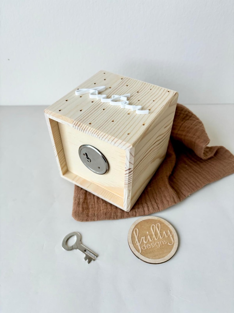 May include: A wooden box with a keyhole and a white inscription on the top. The box is sitting on a brown fabric surface. A key and a wooden circle with the text "frilly designs" are also visible.