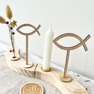 May include: Wooden Advent wreath with four candle holders, two wooden fish symbols, and a small vase for flowers. The wreath is made of natural wood and has a rustic look. The words "frilly designs" are printed on a round wooden tag.