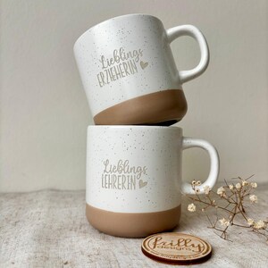 May include: Two ceramic mugs stacked vertically, featuring a white speckled finish with a solid brown base. The top mug displays the text "Lieblings ERZIEHERIN" and the bottom mug displays "Lieblings LEHRERIN", both accompanied by a small heart icon.