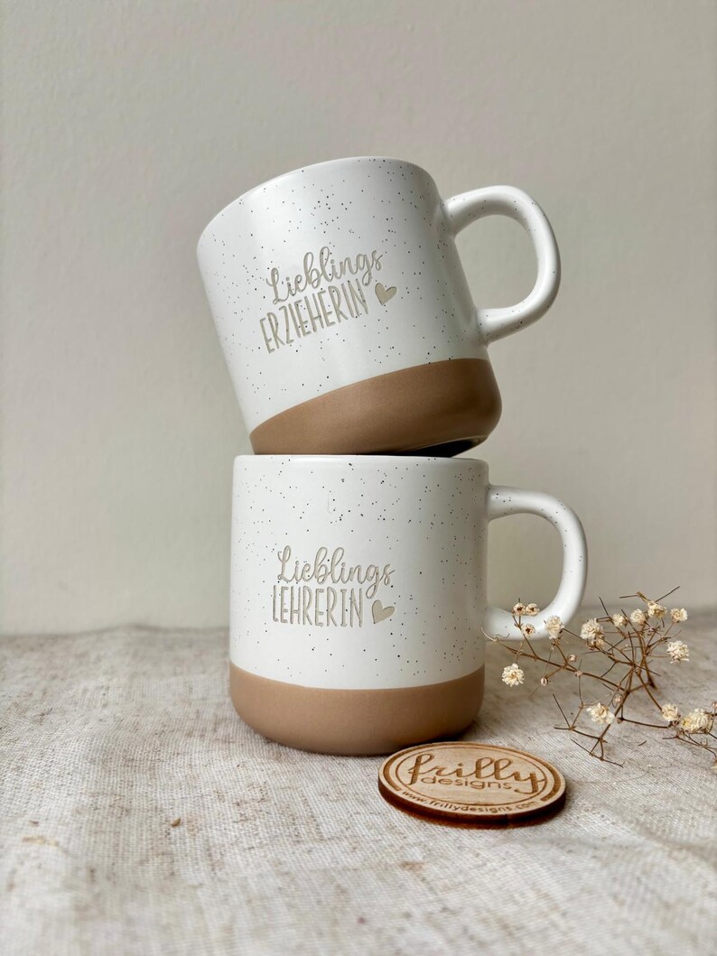 May include: Two ceramic mugs stacked vertically, featuring a white speckled finish with a solid brown base. The top mug displays the text "Lieblings ERZIEHERIN" and the bottom mug displays "Lieblings LEHRERIN", both accompanied by a small heart icon.