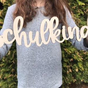 May include: A person holds a wooden sign that says "Schulkind" in cursive lettering. The sign is made of light brown wood and has a natural finish.