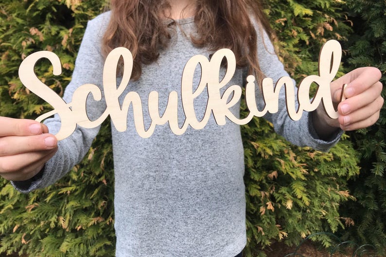 May include: A person holds a wooden sign that says "Schulkind" in cursive lettering. The sign is made of light brown wood and has a natural finish.
