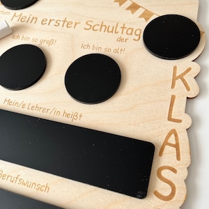 May include: Wooden sign with black chalkboard circles and a rectangular chalkboard. The sign says "Mein erster Schultag" (My first day of school) and "der" (the) and "Ich bin so gro&szlig;!" (I am so big!) and "Ich bin so alt!" (I am so old!) and "Mein/e Lehrer/in hei&szlig;t" (My teacher's name is) and "Berufswunsch" (Career wish). The sign also has the word "KLASS" (Class) written on it.