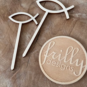 May include: Two wooden cutout fish symbols with a cross in the middle, standing upright on a wooden surface. A round wooden sign with the text "frilly designs" is in the lower right corner of the image.