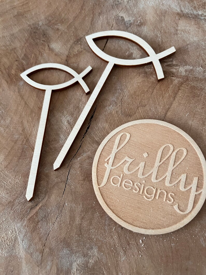 May include: Two wooden cutout fish symbols with a cross in the middle, standing upright on a wooden surface. A round wooden sign with the text "frilly designs" is in the lower right corner of the image.
