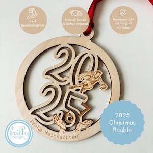 Op de afbeelding: Een houten kerstbal met het jaar 2025 uitgesneden, met een basketballer en de woorden "Frohe Weihnachten". De bal heeft een rood lint om op te hangen en de tekst "2025 Christmas Bauble".