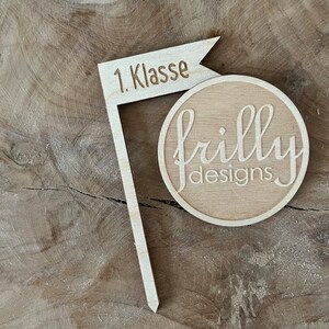 May include: A wooden flag-shaped sign with the text "1. Klasse" and a round wooden piece with the text "frilly designs" in a cursive font. The items are made of light-colored wood and are set against a wood grain background.