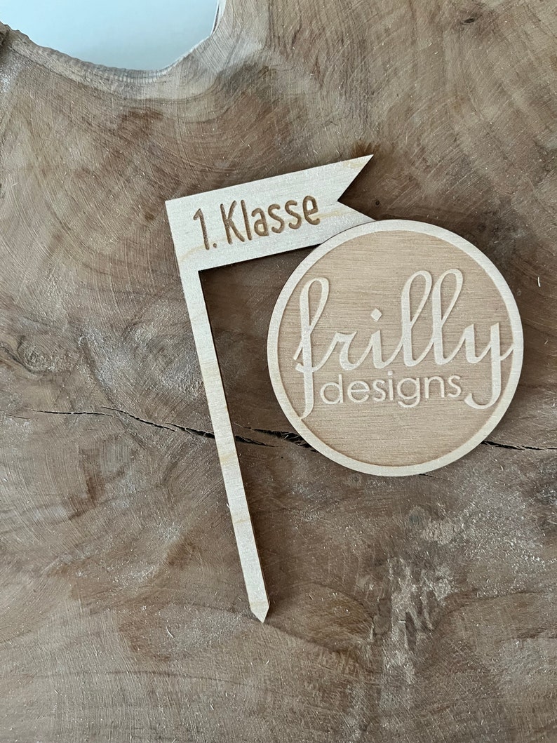 May include: A wooden flag-shaped sign with the text "1. Klasse" and a round wooden piece with the text "frilly designs" in a cursive font. The items are made of light-colored wood and are set against a wood grain background.