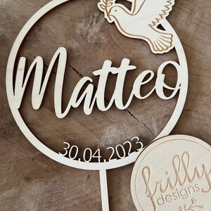 May include: A wooden circle with the name Matteo and the date 30.04.2023 engraved on it. A dove with an olive branch is on the top of the circle. The circle is attached to a wooden stick. A round wooden tag with the text "frilly designs" is in the bottom right corner of the image.