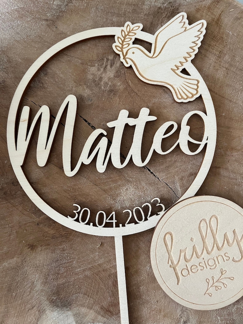 May include: A wooden circle with the name Matteo and the date 30.04.2023 engraved on it. A dove with an olive branch is on the top of the circle. The circle is attached to a wooden stick. A round wooden tag with the text "frilly designs" is in the bottom right corner of the image.