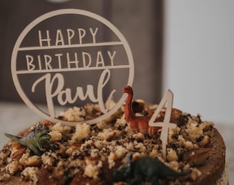 May include: A chocolate cake decorated with a wooden sign that says "Happy Birthday Paul" and a number four. The cake is also decorated with dinosaur figurines and chocolate crumbs.