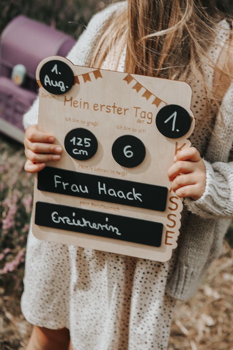 May include: A wooden sign with a chalkboard background and a black and white banner design. The sign says "Mein erster Tag" (My first day) and includes the date "1. Aug." (August 1st), the child's height "125 cm", age "6", teacher's name "Frau Haack", and profession "Erzieherin" (Educator).