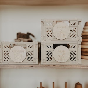 Könnte beinhalten: Drei hellfarbene Holz-Aufbewahrungskisten mit runden Etiketten. Auf einem Etikett steht "Kuscheltiere" mit einer Bärenillustration, auf einem anderen "Bausteine" mit Buchstaben und auf dem dritten "Figuren" mit einem Schaukelpferd. Die Kisten sind gestapelt.