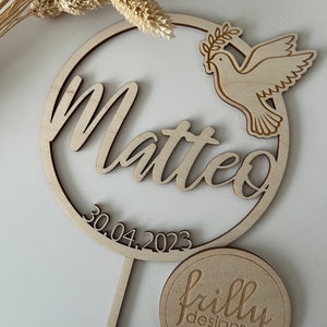 May include: Wooden cake topper with a circle design featuring the name Matteo and the date 30.04.2023. A dove with an olive branch is on the right side of the circle. The topper is on a wooden stick. A round wooden tag with the text "frilly designs" is in the bottom right corner of the image.