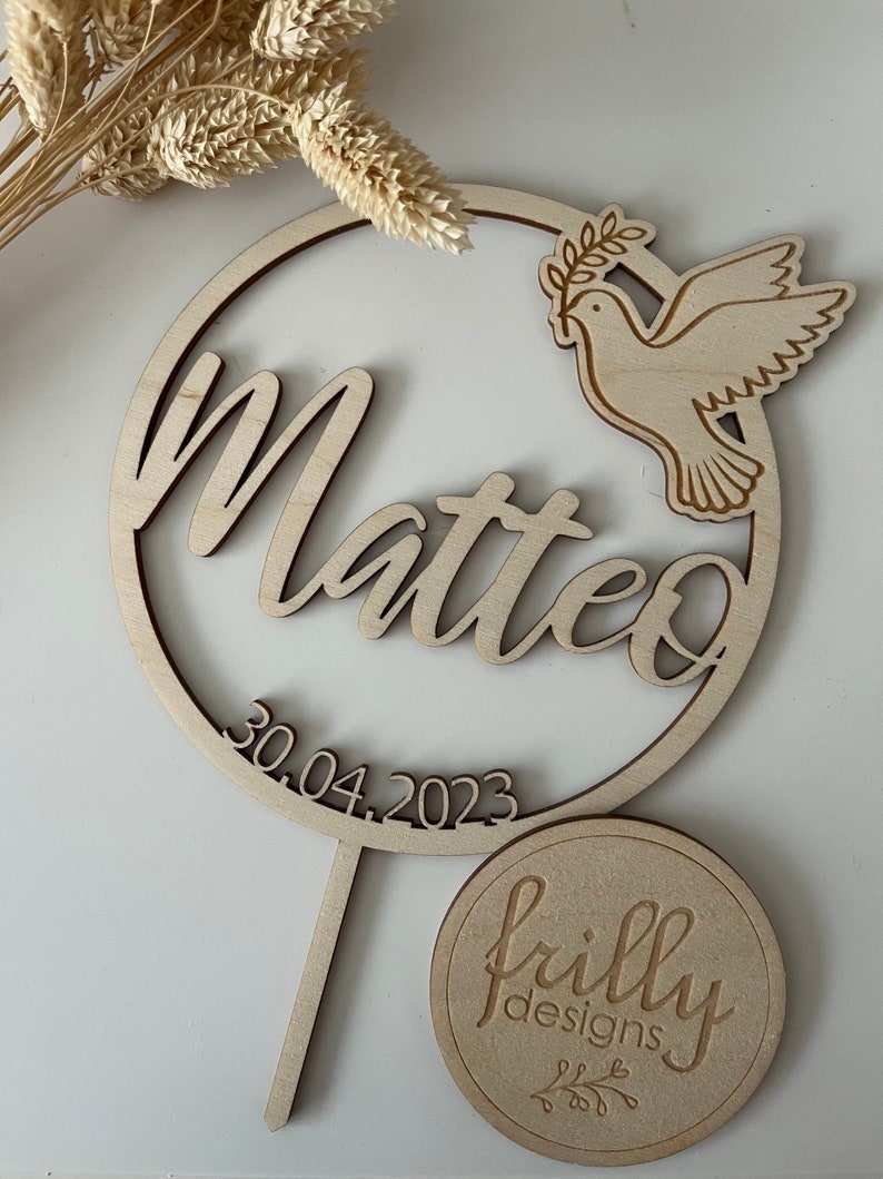 May include: Wooden cake topper with a circle design featuring the name Matteo and the date 30.04.2023. A dove with an olive branch is on the right side of the circle. The topper is on a wooden stick. A round wooden tag with the text "frilly designs" is in the bottom right corner of the image.