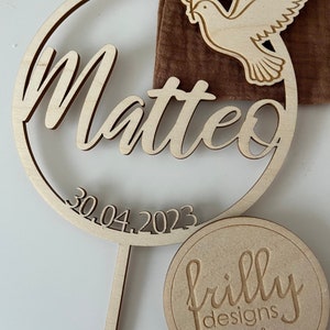 May include: Wooden cake topper with a circle design featuring the name Matteo and the date 30.04.2023. A dove with an olive branch is on the top of the circle. The topper is on a wooden stick. A round wooden tag with the text "frilly designs" is in the background.