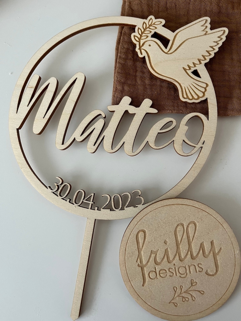 May include: Wooden cake topper with a circle design featuring the name Matteo and the date 30.04.2023. A dove with an olive branch is on the top of the circle. The topper is on a wooden stick. A round wooden tag with the text "frilly designs" is in the background.