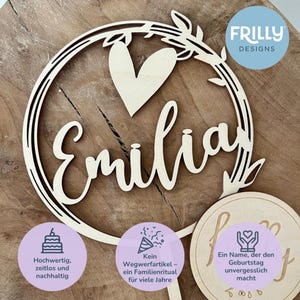 May include: A circular wooden cake topper featuring the name Emilia in a cursive script, topped with a heart cutout and a partial leafy branch border. The design is light-colored wood with dark laser-cut edges, presented as a decorative piece.