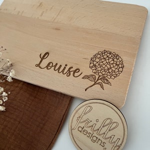 Peut inclure: Une planche &agrave; d&eacute;couper en bois clair grav&eacute;e du nom "Louise" et d'un motif floral. Un petit dessous de verre en bois avec le texte "frilly designs" est &eacute;galement visible.