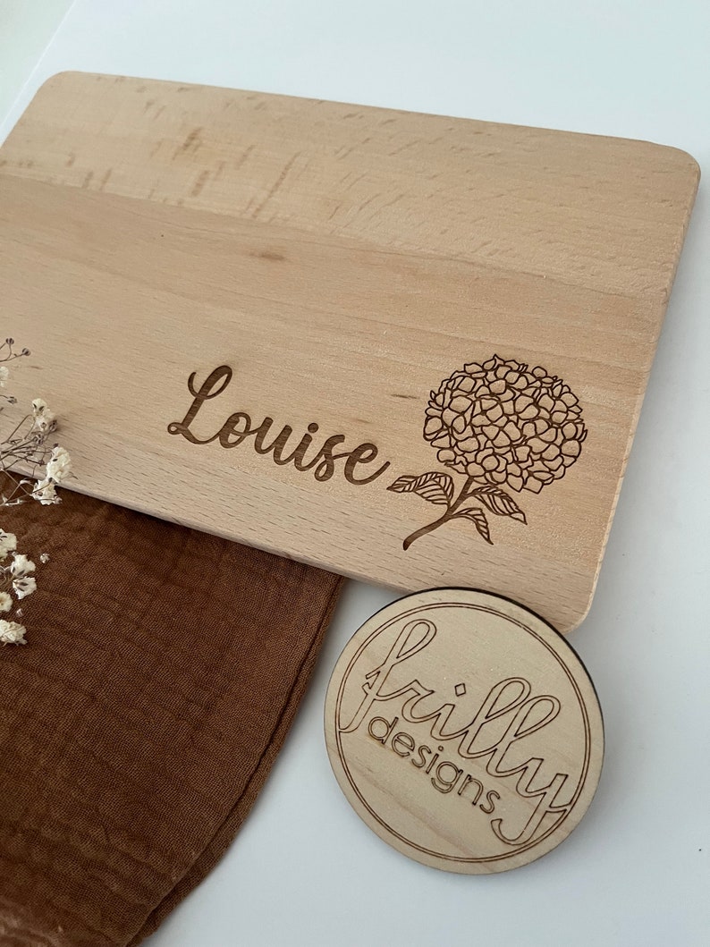 Peut inclure: Une planche &agrave; d&eacute;couper en bois clair grav&eacute;e du nom "Louise" et d'un motif floral. Un petit dessous de verre en bois avec le texte "frilly designs" est &eacute;galement visible.