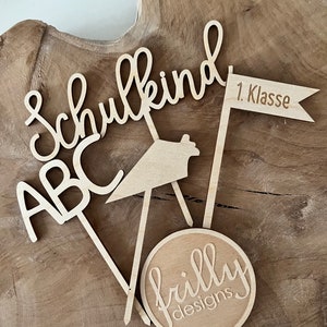May include: Wooden cake toppers with the words "Schulkind", "ABC", and "1. Klasse" on a flag. A circular topper with "frilly designs" is also included. The toppers are made of light-colored wood and are arranged on a wooden surface.