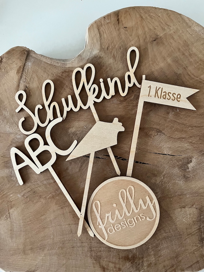 May include: Wooden cake toppers with the words "Schulkind", "ABC", and "1. Klasse" on a flag. A circular topper with "frilly designs" is also included. The toppers are made of light-colored wood and are arranged on a wooden surface.