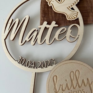 May include: Wooden circle with the name Matteo and the date 30.04.2023. A dove with an olive branch is in the top right corner. The bottom of the circle has a stick attached. A small wooden circle with the text "frilly designs" is in the bottom right corner.