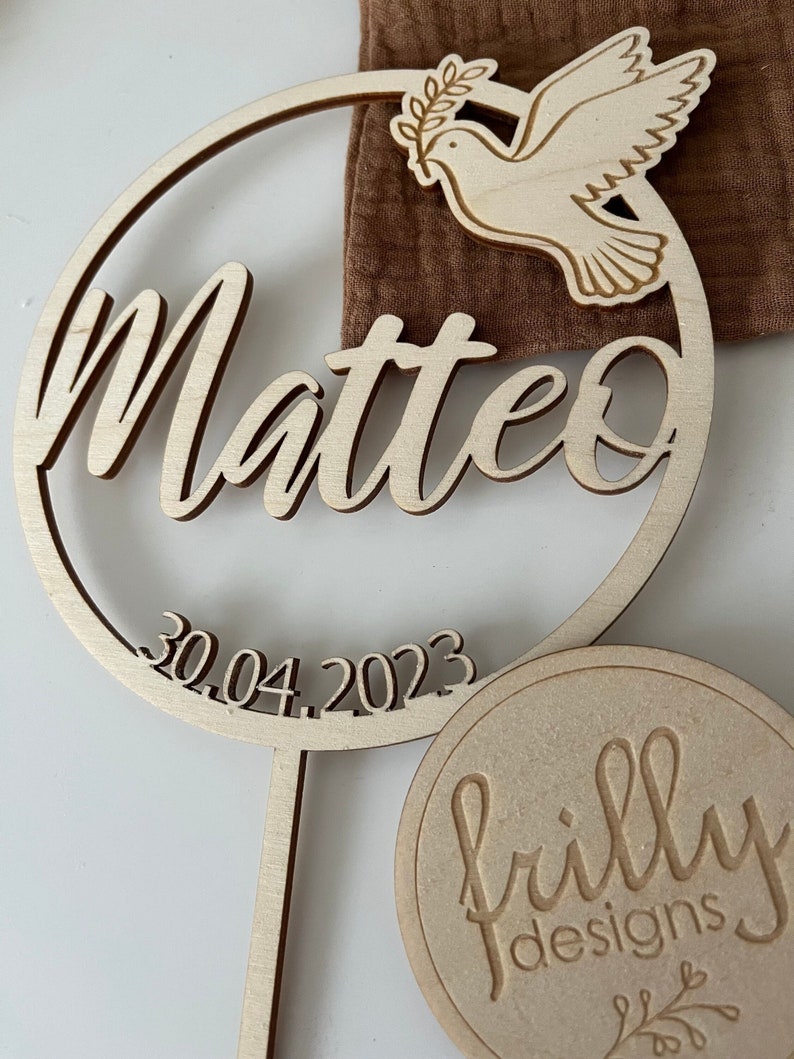 May include: Wooden circle with the name Matteo and the date 30.04.2023. A dove with an olive branch is in the top right corner. The bottom of the circle has a stick attached. A small wooden circle with the text "frilly designs" is in the bottom right corner.