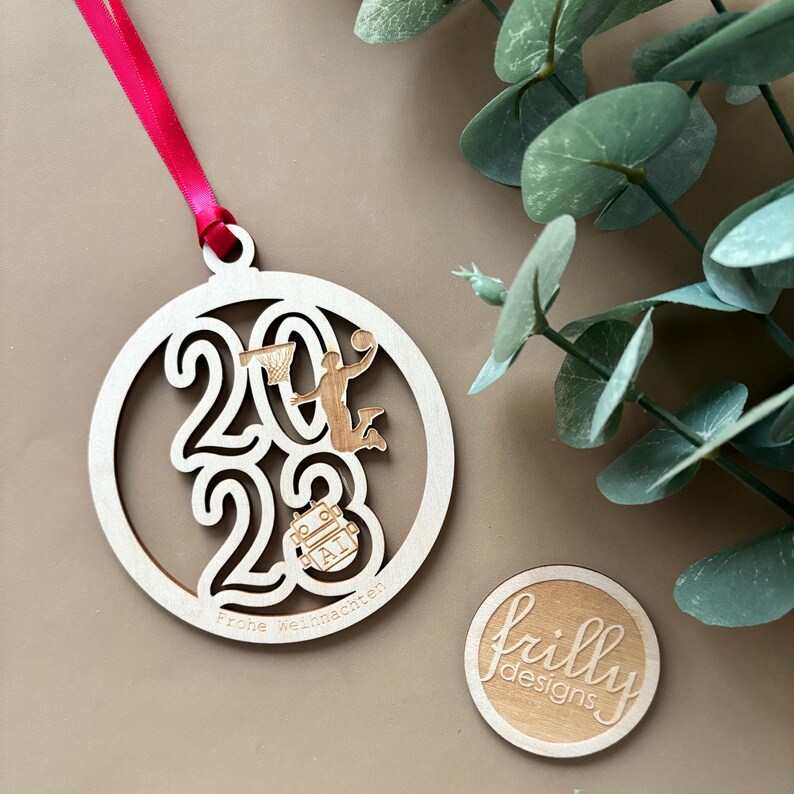 Op de afbeelding: Een houten kerstversiering met het jaar 2023 uitgesneden, met een basketballer en de woorden "Frohe Weihnachten". De versiering is rond en heeft een rood lint om op te hangen. Een kleine houten cirkel met de tekst "frilly designs" is ook zichtbaar.