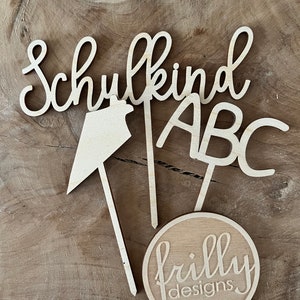 May include: Wooden cake toppers with the words "Schulkind", "ABC", and a house shape. A round topper with "frilly designs" is also included. The toppers are light brown and sit on a wooden surface.