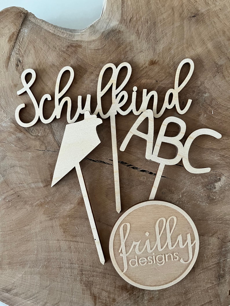 May include: Wooden cake toppers with the words "Schulkind", "ABC", and a house shape. A round topper with "frilly designs" is also included. The toppers are light brown and sit on a wooden surface.