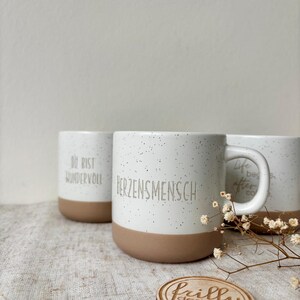May include: Three ceramic mugs with a speckled white finish and a solid tan base. The front mug features the text "HERZENSMENSCH" in gray. The left mug displays "DU BIST WUNDERVOLL" and the right mug shows "Life begins after coffee" in gray.