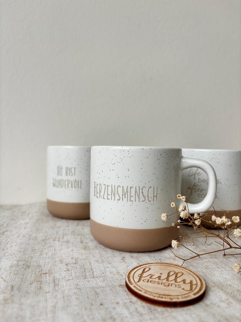 May include: Three ceramic mugs with a speckled white finish and a solid tan base. The front mug features the text "HERZENSMENSCH" in gray. The left mug displays "DU BIST WUNDERVOLL" and the right mug shows "Life begins after coffee" in gray.