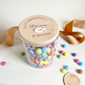 May include: A glass jar filled with colorful candy, with a wooden lid that says "Zur Kommunion Marcus 29. April 2023" and a white fish symbol.