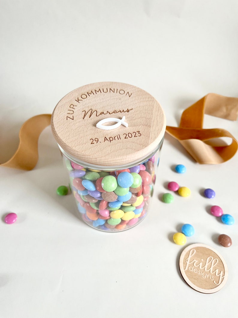 May include: A glass jar filled with colorful candy, with a wooden lid that says "Zur Kommunion Marcus 29. April 2023" and a white fish symbol.