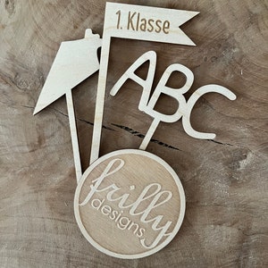May include: A wooden school-themed decoration with the text "1. Klasse" on a flag, "ABC" letters, and a circular logo that says "frilly designs". The decoration is made of light-colored wood.