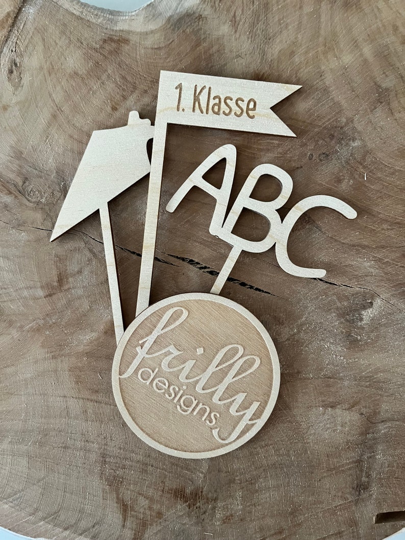 May include: A wooden school-themed decoration with the text "1. Klasse" on a flag, "ABC" letters, and a circular logo that says "frilly designs". The decoration is made of light-colored wood.