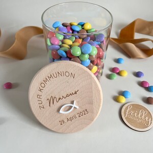 May include: A wooden jar lid with the text "Zur Kommunion Marcus 29. April 2023" and a small white fish symbol. The lid is on top of a glass jar filled with colorful candy.