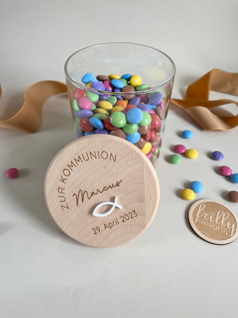 May include: A wooden jar lid with the text "Zur Kommunion Marcus 29. April 2023" and a small white fish symbol. The lid is on top of a glass jar filled with colorful candy.