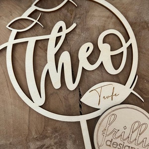 May include: Wooden cake topper featuring the name "Theo" in a cursive script, encircled by a circle with leaf motifs. The topper also includes the word "Taufe" and a small circle with the text "frilly designs".
