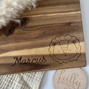 May include: Personalized wooden cutting board with the name "Marcus" engraved on it. The board features a cute lion design and a small round wooden tag with the text "frilly designs".