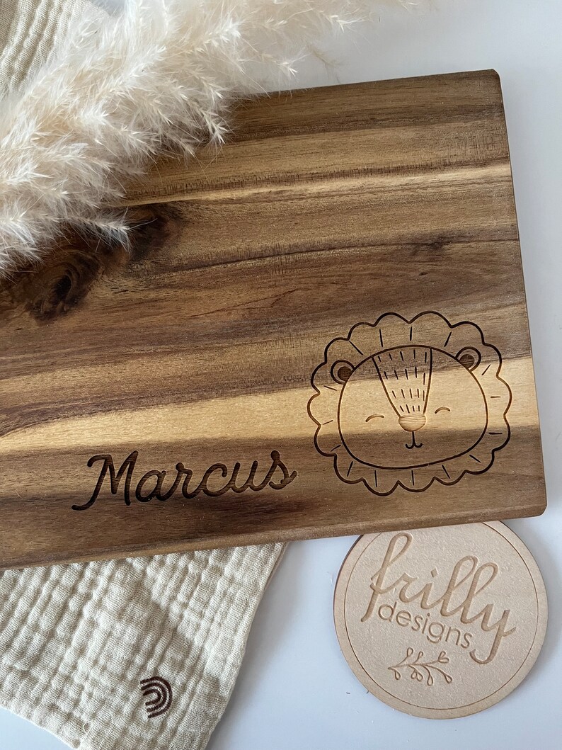 May include: Personalized wooden cutting board with the name "Marcus" engraved on it. The board features a cute lion design and a small round wooden tag with the text "frilly designs".