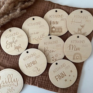May include: A set of ten wooden round ornaments with personalized names and the year 2023 engraved on them. Each ornament features a small graphic design, such as a deer, a rocket, a dinosaur, a fire truck, and a rainbow. The ornaments are perfect for a baby shower or a birthday party.