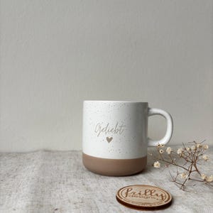 May include: A white ceramic mug with a speckled finish and a solid brown base. The front of the mug features the word Geliebt in a cursive script font above a small heart icon. A small round wooden disc with the text Frilly Designs is nearby.