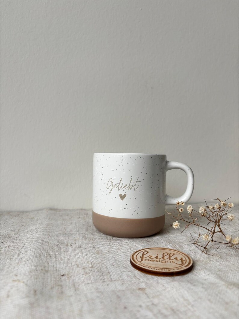 May include: A white ceramic mug with a speckled finish and a solid brown base. The front of the mug features the word Geliebt in a cursive script font above a small heart icon. A small round wooden disc with the text Frilly Designs is nearby.