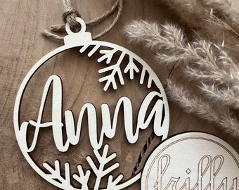 May include: A wooden Christmas bauble with the name "Anna" cut out in a script font, surrounded by snowflake designs. The bauble is suspended by a twine loop. A small wooden disc with "frilly designs" is also visible.